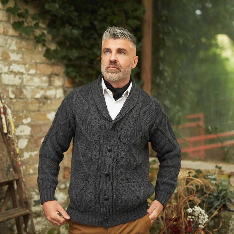 Man standing in an outdoor garden wearing a charcoal colour aran knitted cardigan
